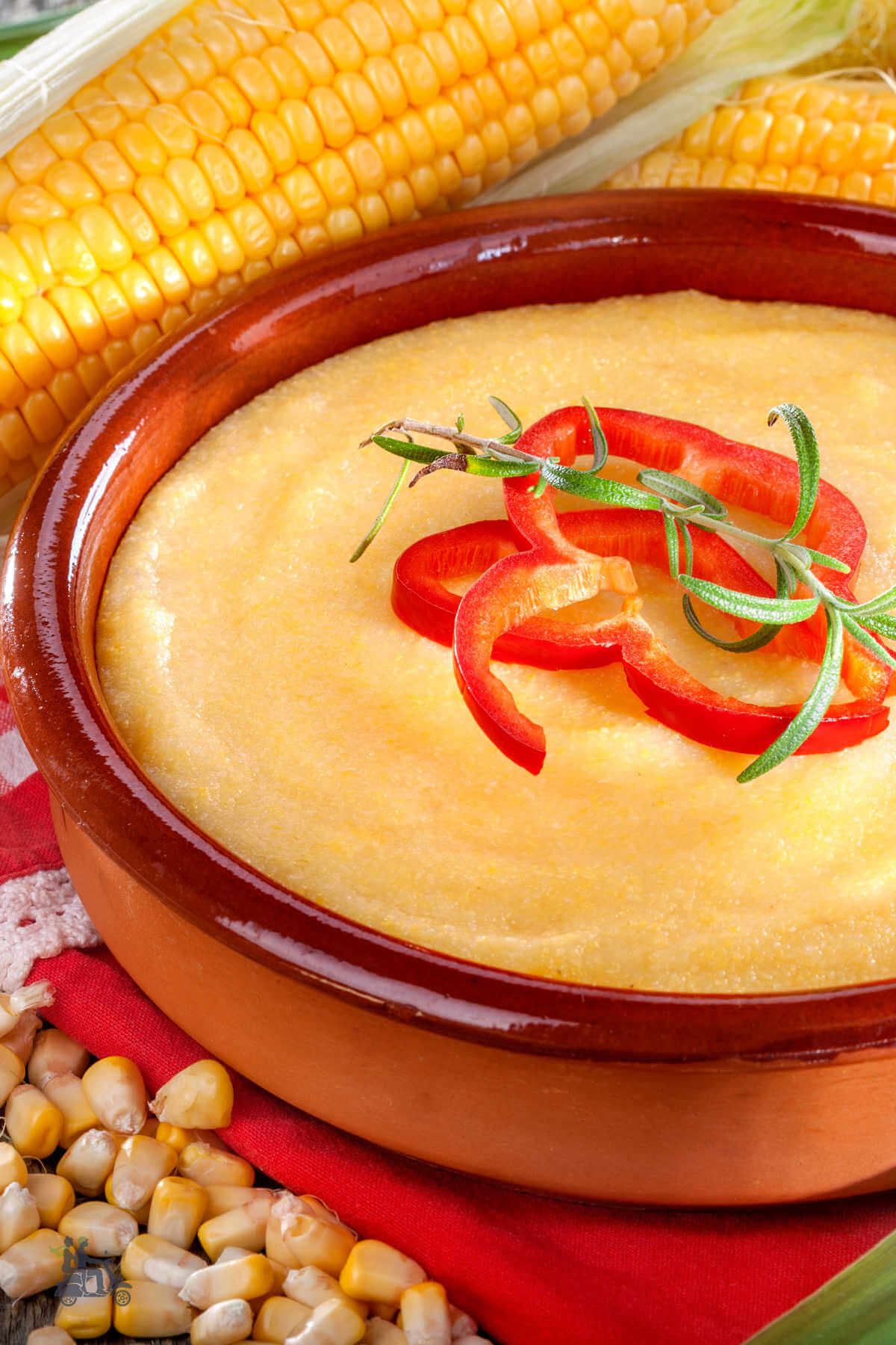 A brown Pottery Bowl filled with cooked creamy cheesy yellow corn grits and ears of yellow corn in the background. 