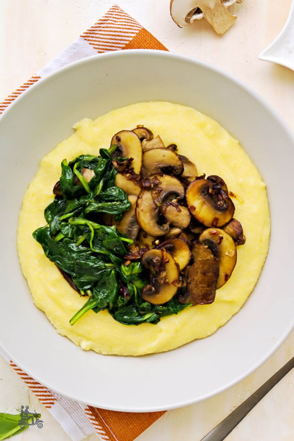 A white plate filled with a serving of cooked cheesy grits topped with cooked wilted spinach and sautéd mushrooms. 