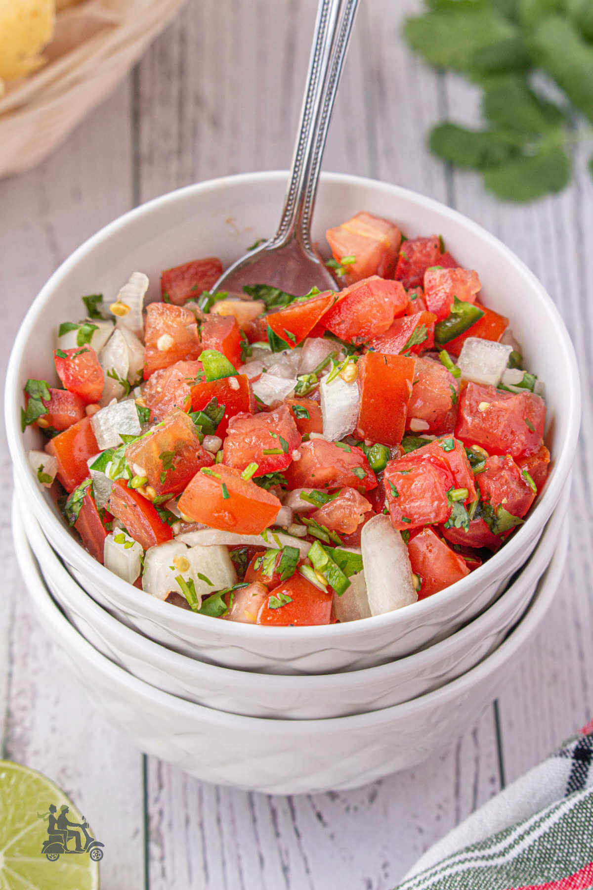 A white bowl filled with a fresh tomato, onion, cilantro mixed salsa with tortilla chips in the background. 