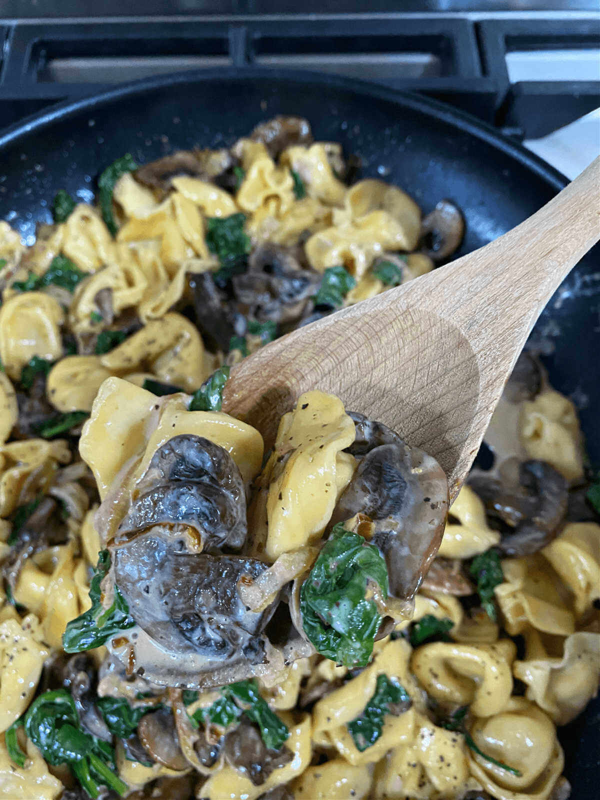 A wooden spoon filled with cheese tortellini, caramelized onions, spinach and mushrooms.