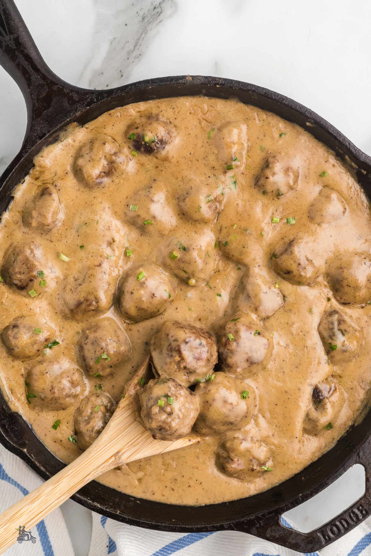 Black Iron Skillet with the Swedish meatballs covered in a rich brown gravy and a wooden spoon dipping into the pan and lifting three meatballs. 