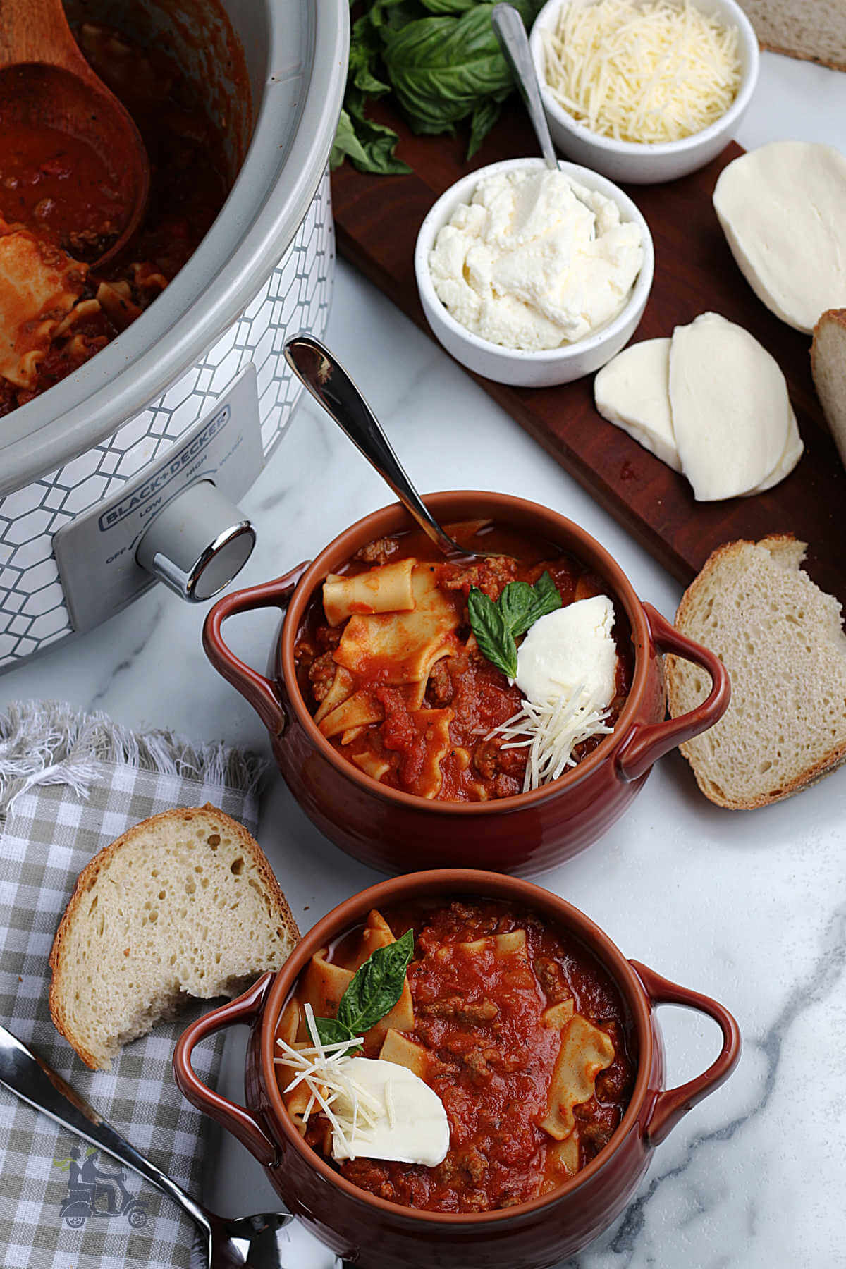 Overhead view of two brown earthenware bowls holding red lasagna soup and topped with parmesan, fresh mozzarella, and a basil sprig. 