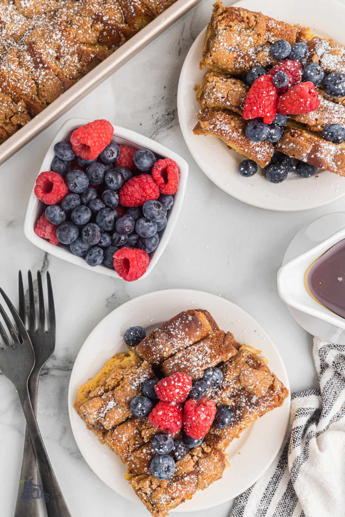 French toast casserole with a serving on a white plate and the dish topped with blueberries and raspberries. 