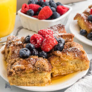 Serving of Brioche French Toast Casserole topped with powdered sugar and maple syrup with fresh raspberries and blueberries.