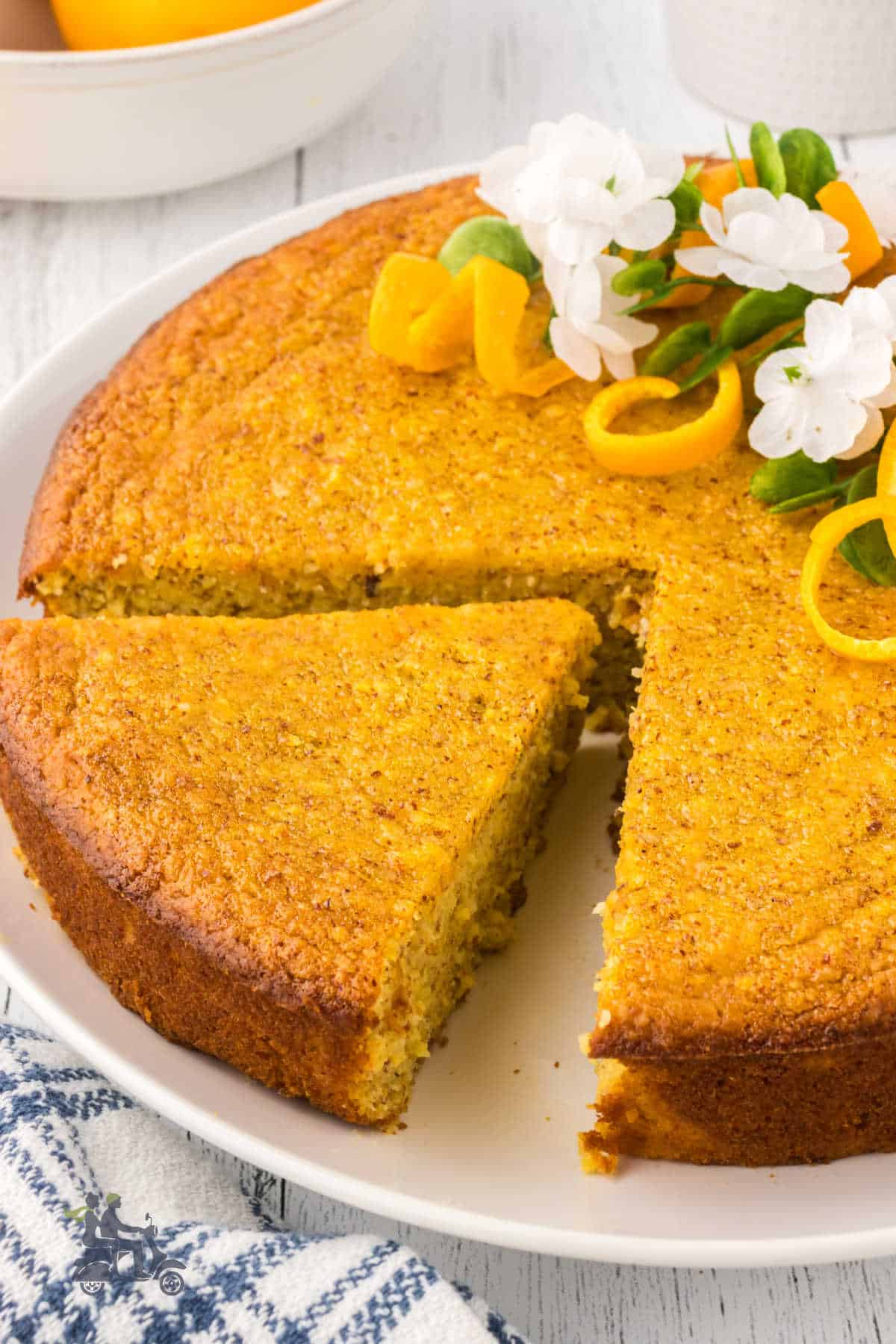 A slice is cut from a round orange almond cake that's on a white plate. 