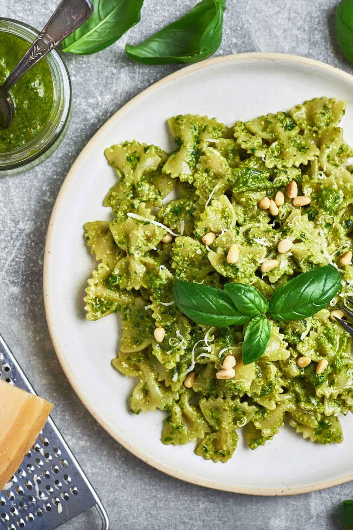 A white plate filled with farfalle pasta al pesto with a sprig of basil on top and sprinkled with pine nuts. 