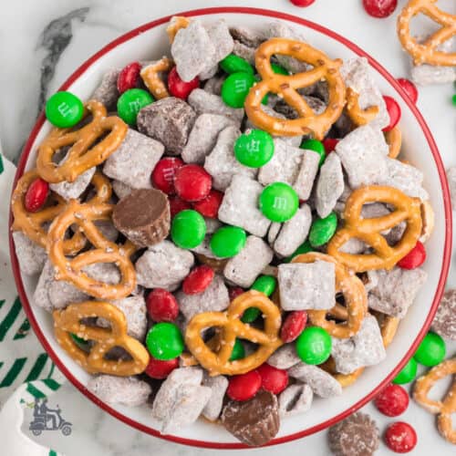 A red bowl filled with sweetened Chex cereal mix with pretzels, M and M candies, Reese's cups, and dusted with powdered sugar.