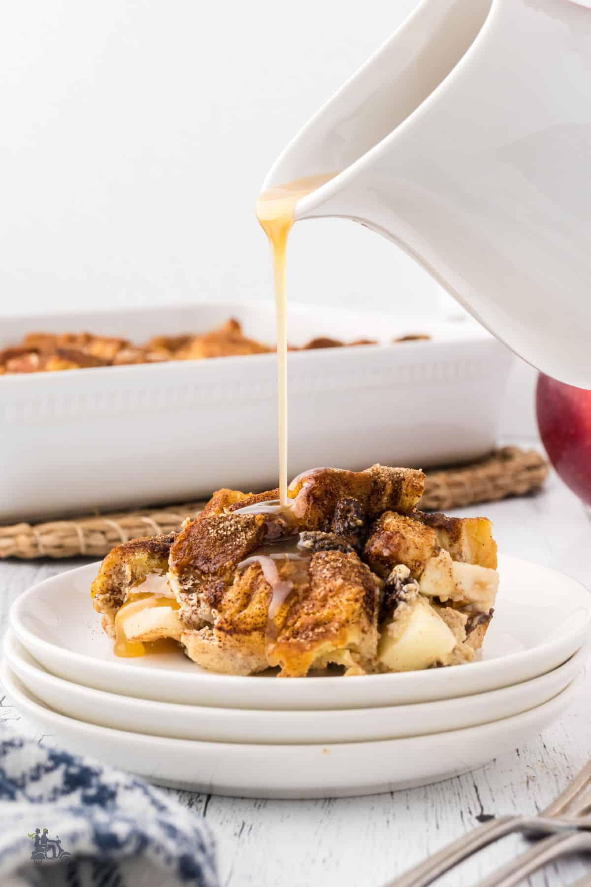 Apple Pie Brioche Bread Pudding on a white plate with a drizzle of Caramel Bourbon Sauce being poured over it. 