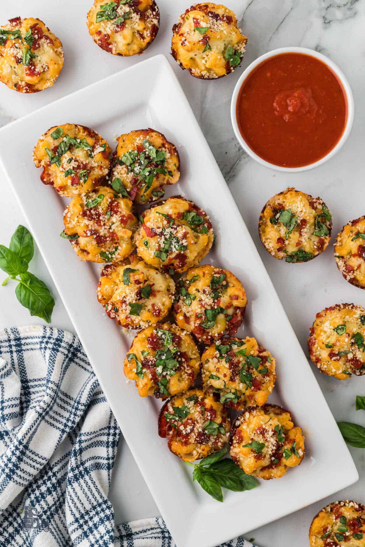 A white rectangular platter holding appetizer pizza bites with more pizza pasta muffins on the table with a bowl of marinara dipping sauce. 