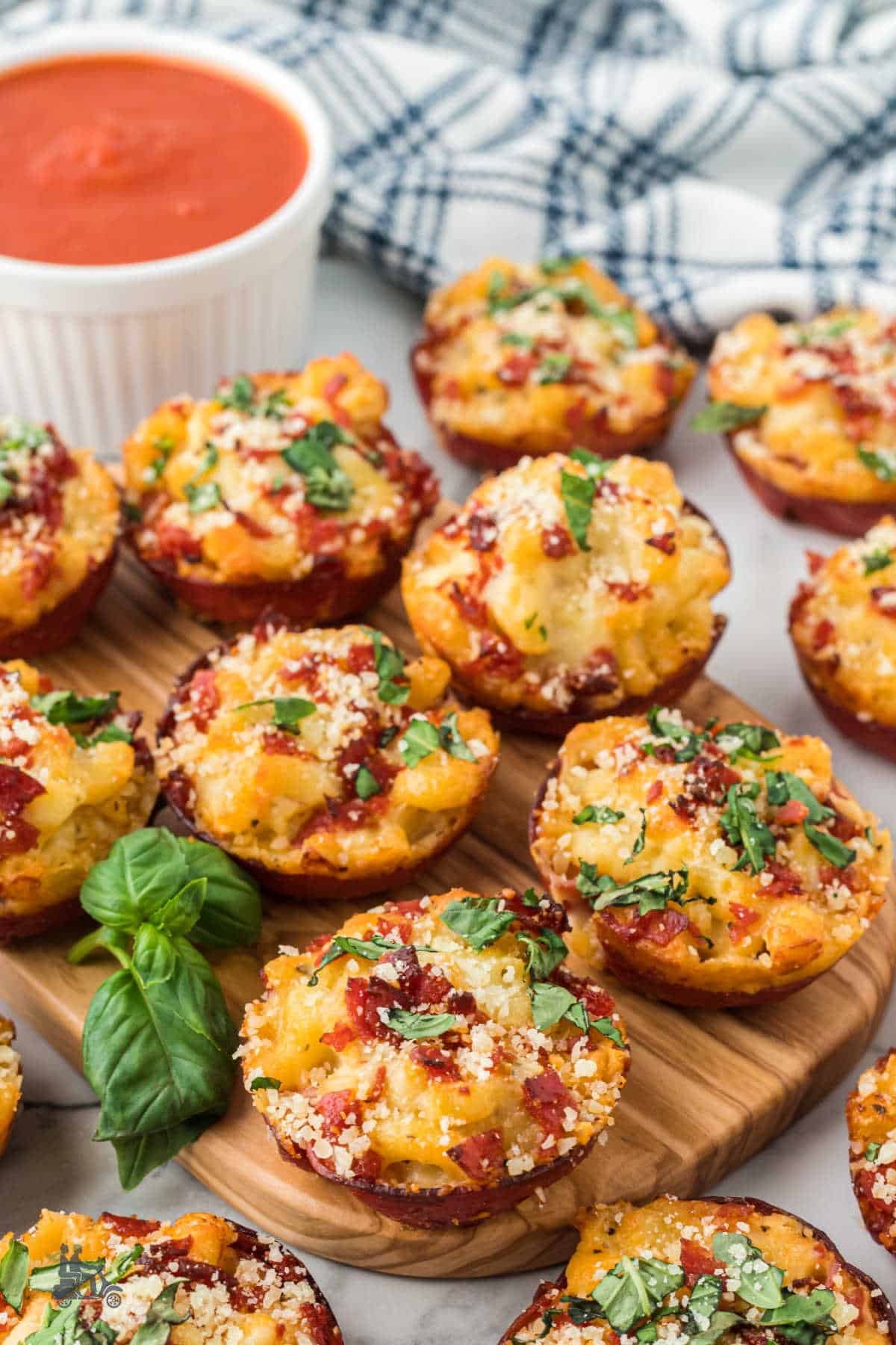 Macaroni and Cheese Pizza Bites on a wood cutting board with a white bowl of tomato marinara on the side. 