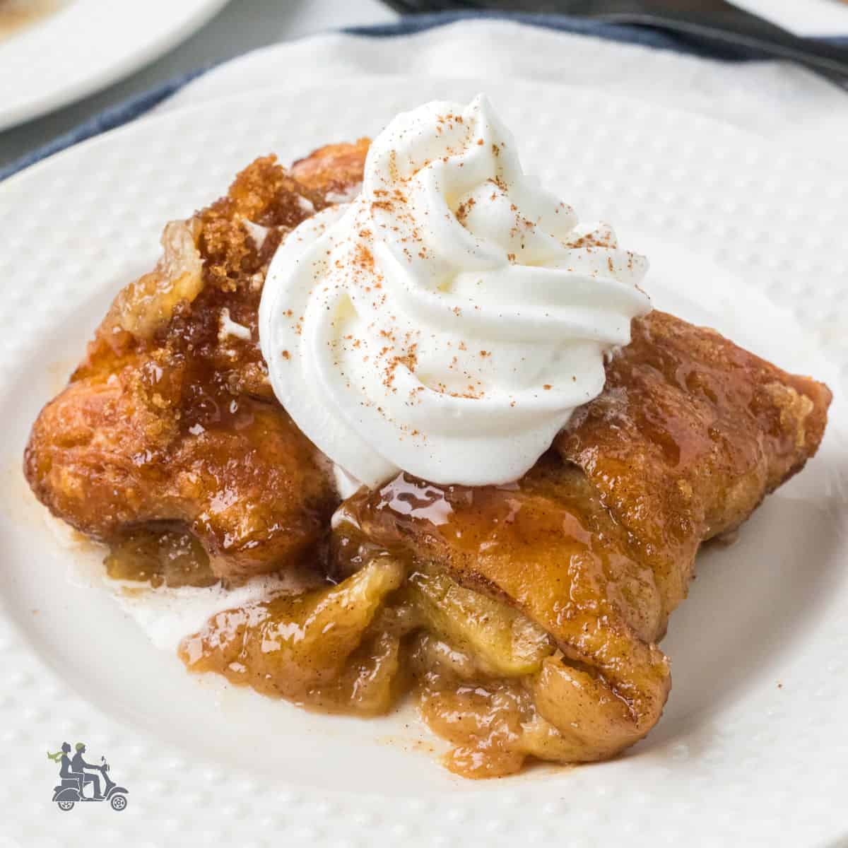 Apples rolled in crescent rolls, baked, and topped with a caramel sauce are served on a white plate with a dollop of whipped cream on top.