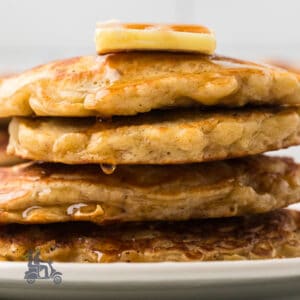 A stack of oatmeal pancakes with a pat of butter on top and maple syrup dripping down the stack.