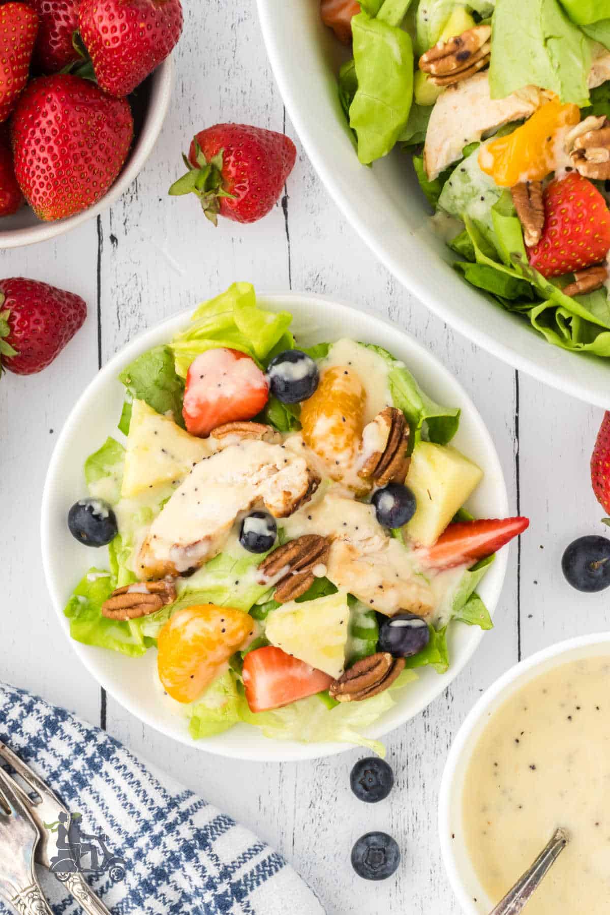 A bowl of Chicken Strawberry Poppyseed salad is on a white wooden table. There are more strawberries on one side and the serving bowl with the salad on the other. 