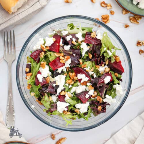 A glass bowl filled with roasted sliced beets with goat cheese, mesclun salad, and toasted walnuts.