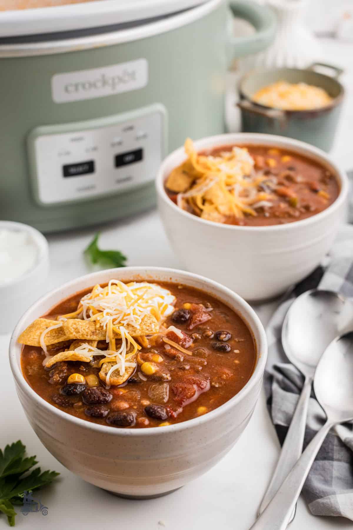 Two bowls of tomato-rich taco soup with sour cream and corn chip toppings, with the crockpot in the background. 