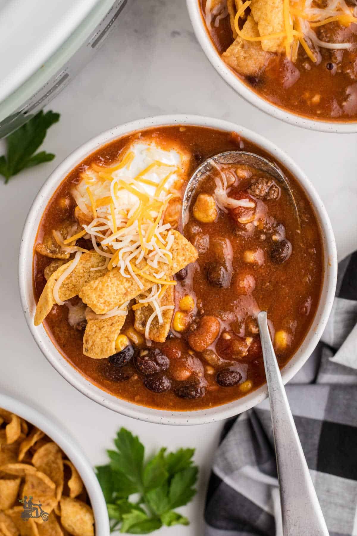 A white bowl filled with taco soup made in a crockpot and garnished with sour cream, corn chips, and shredded cheddar cheese. 