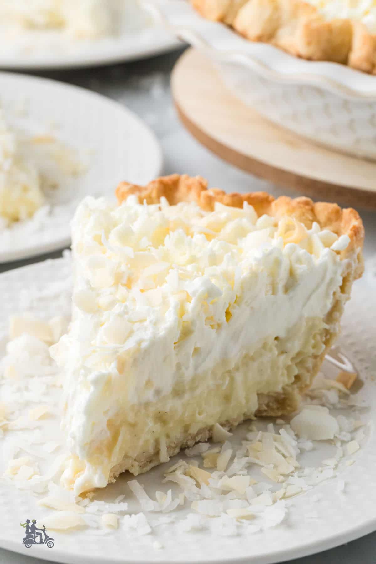 A slice of triple cream coconut pie on a white China plate with coconut sprinkled around it.