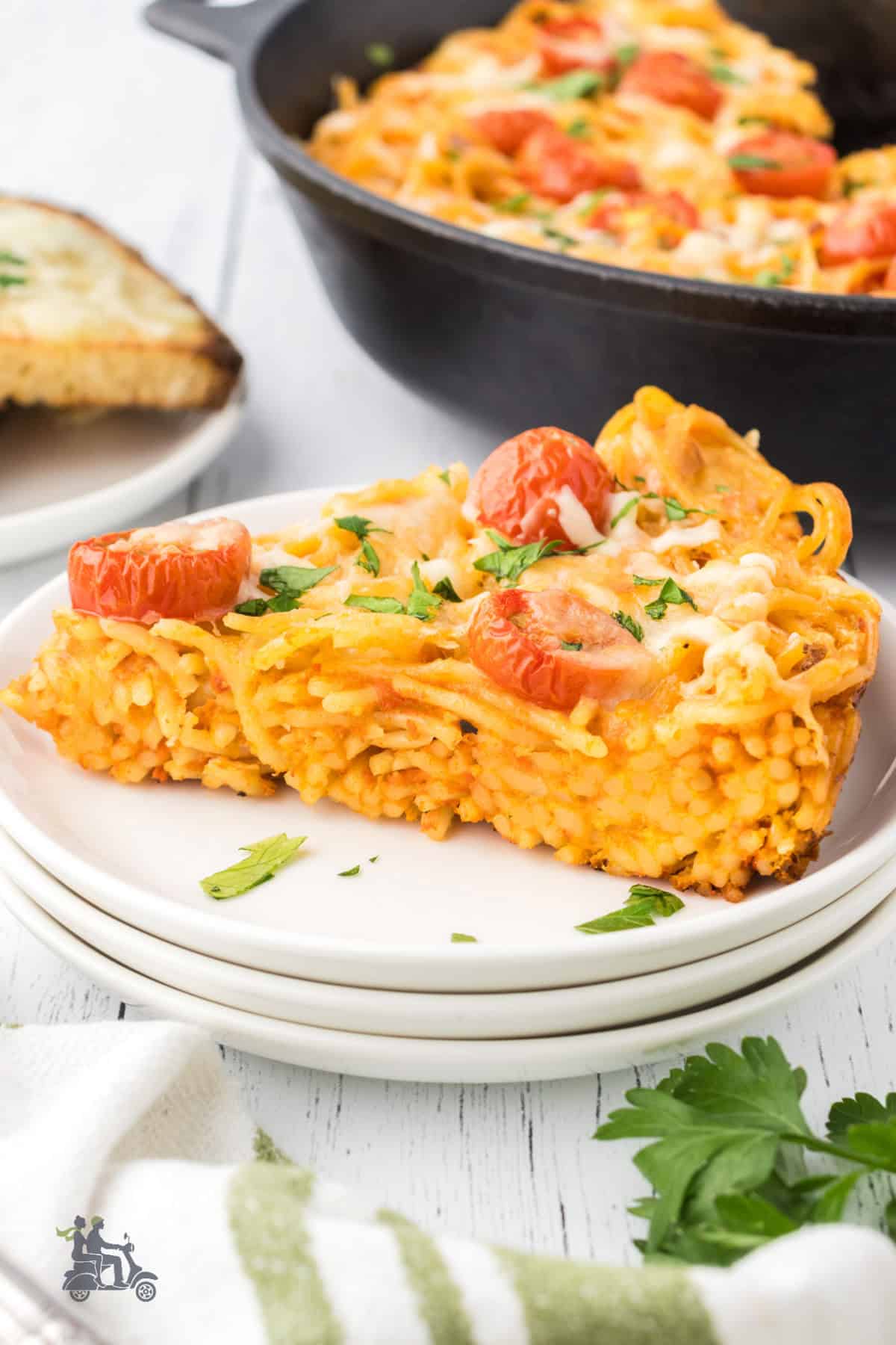 A wedge of the spaghetti frittata is placed on a white plate with the black cast iron skillet and the rest of the frittata in the background. 
