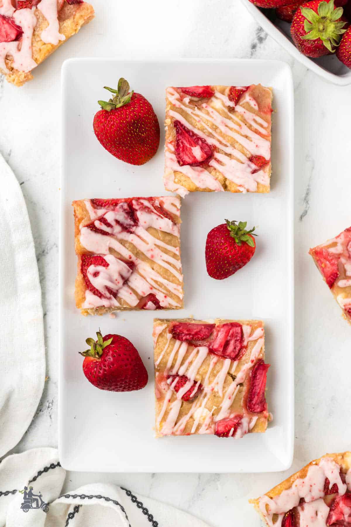 Three White Chocolate Blondies made with strawberries are on a white serving platter with loose fresh strawberries on the plate. 