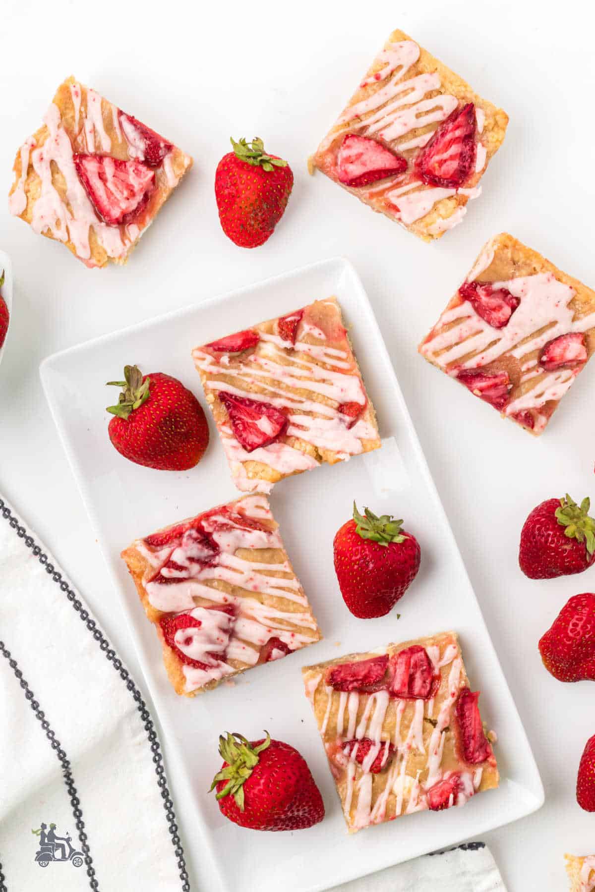 Strawberry Blondies with White Chocolate are on a white platter with fresh strawberries on the platter and table.