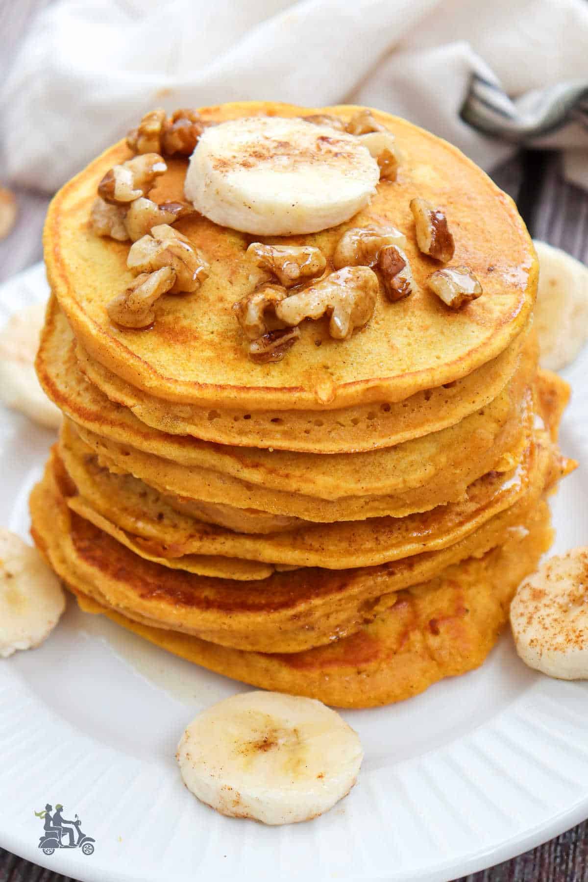 The finished cooked banana pumpkin pancakes are stacked on a white plate with nuts and banana slices ready for the maple syrup finish. 