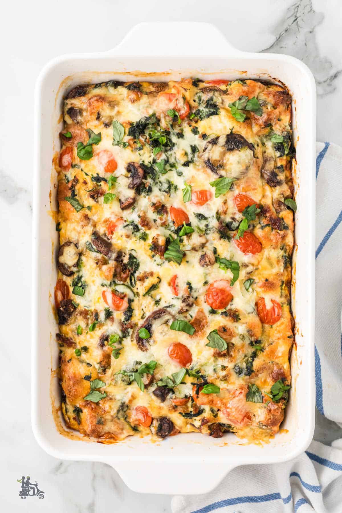 A white casserole dish with a baked breakfast strata made with sausage, spinach, mushrooms, and tomatoes. 