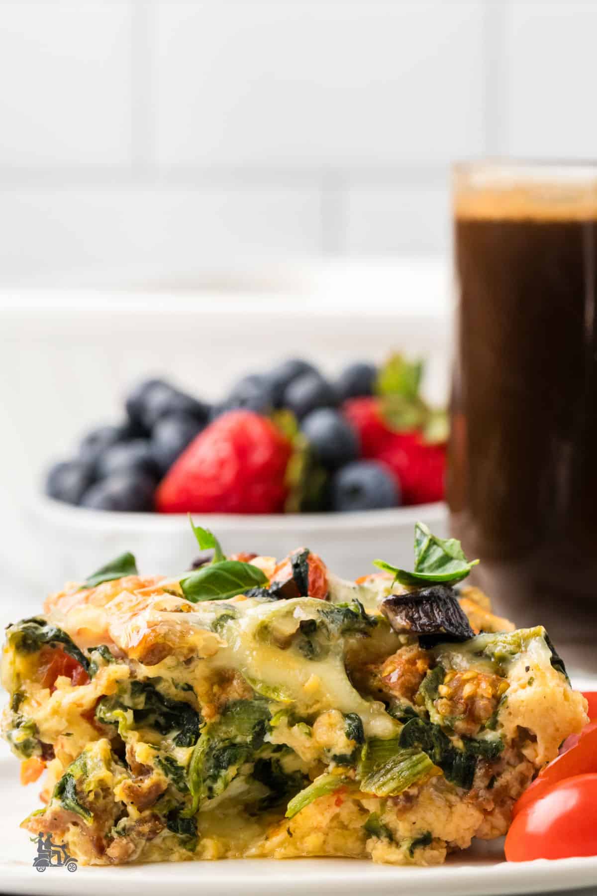 Side view of the sausage strata on a white plate with black coffee in a glass mug and berries in a bowl. 