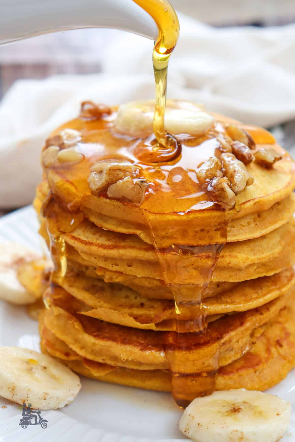 A stack of pumpkin banana pancakes on a white plate with maple syrup poured over them. 