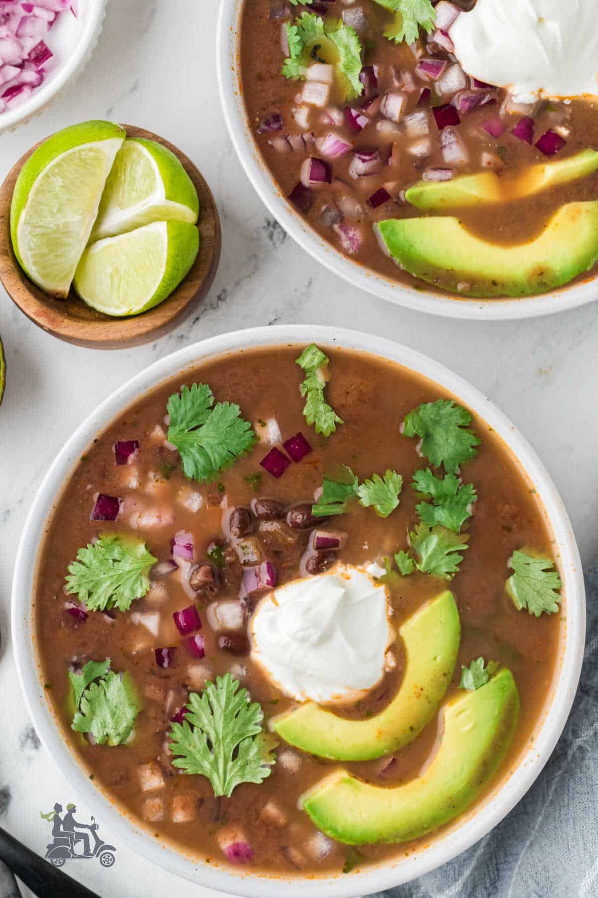 Two soup bowls filled with black bean soup garnished with diced onion, sour cream, sliced avocados, and cilantro leaves. 