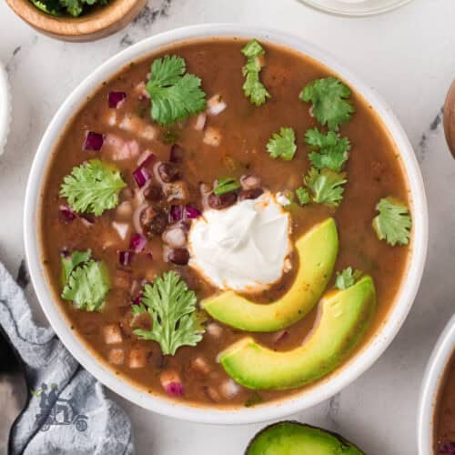 Black Bean soup garnished with sliced avocado, diced red onions, and cilantro leaves.