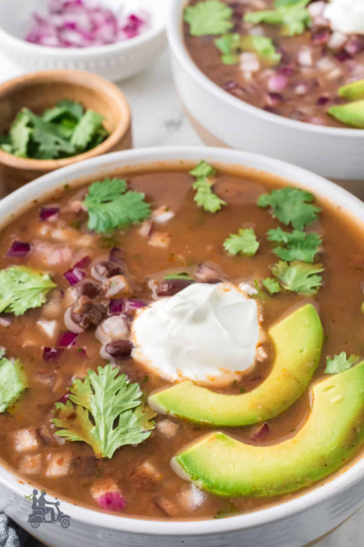 Southwestern-flavored black bean soup is served in a white bowl and garnished with avocado slices, diced red onion, and fresh cilantro leaves. 