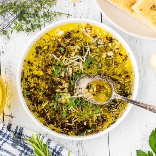 A dipping bowl filled with a garlic herb olive oil bread dip with a spoon in the dip.