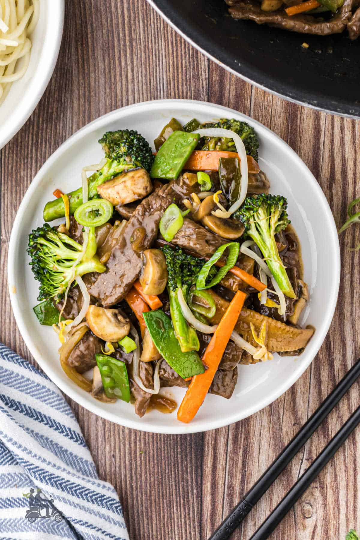 A bowl of chop suey with beef on a wood table with chopsticks on the side. 