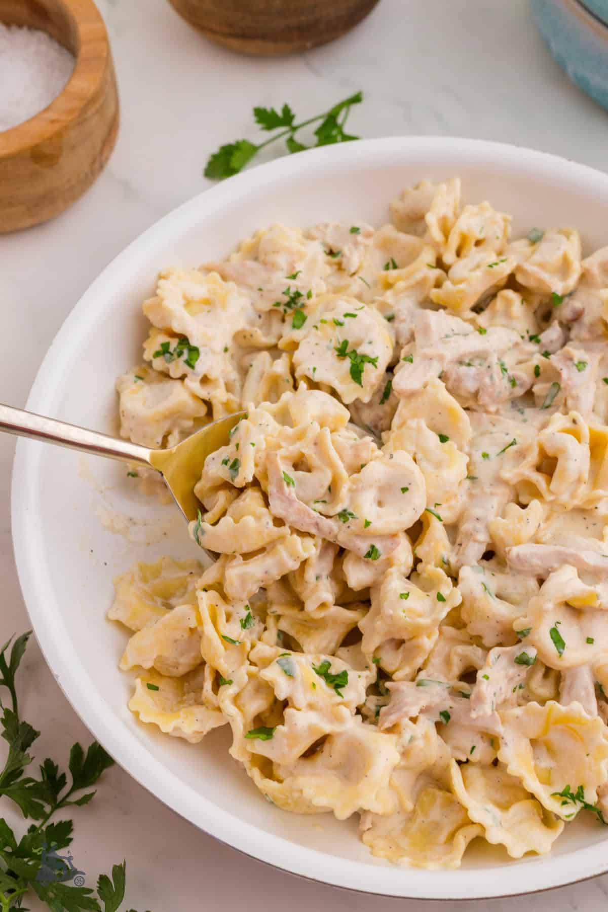 A serving spoon removes a Creamy Alfredo Chicken Tortellini scoop from a white skillet. 