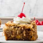 Side view of a square of classic fruit cocktail cake with a dollop of whipped cream and a maraschino cherry on top.