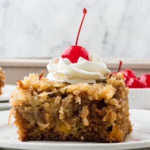 Side view of a square of classic fruit cocktail cake with a dollop of whipped cream and a maraschino cherry on top.