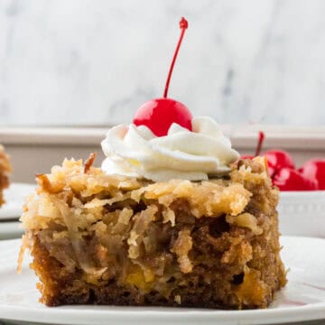 Side view of a square of classic fruit cocktail cake with a dollop of whipped cream and a maraschino cherry on top.
