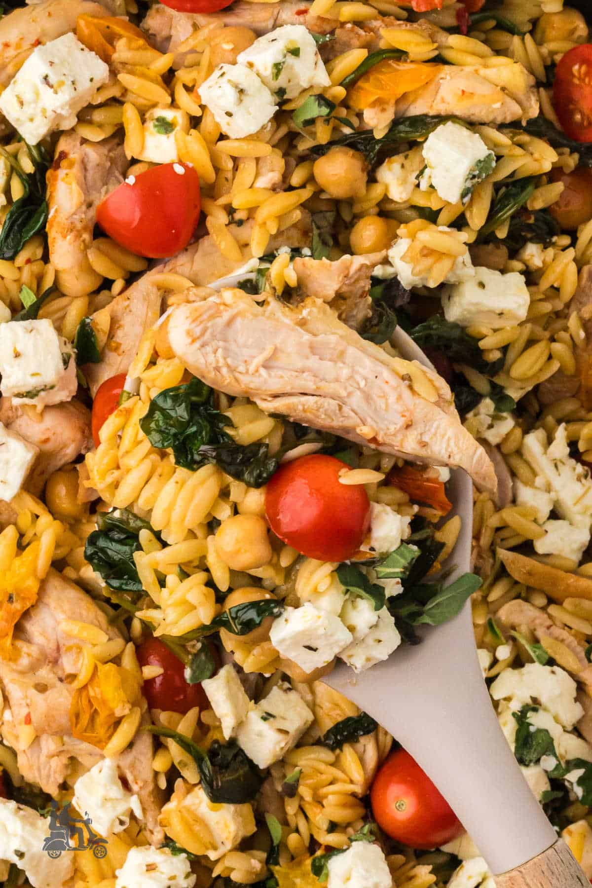 Close-up of a wooden spoon holding a scoop of chicken, pasta, spinach, and feta cheese.  