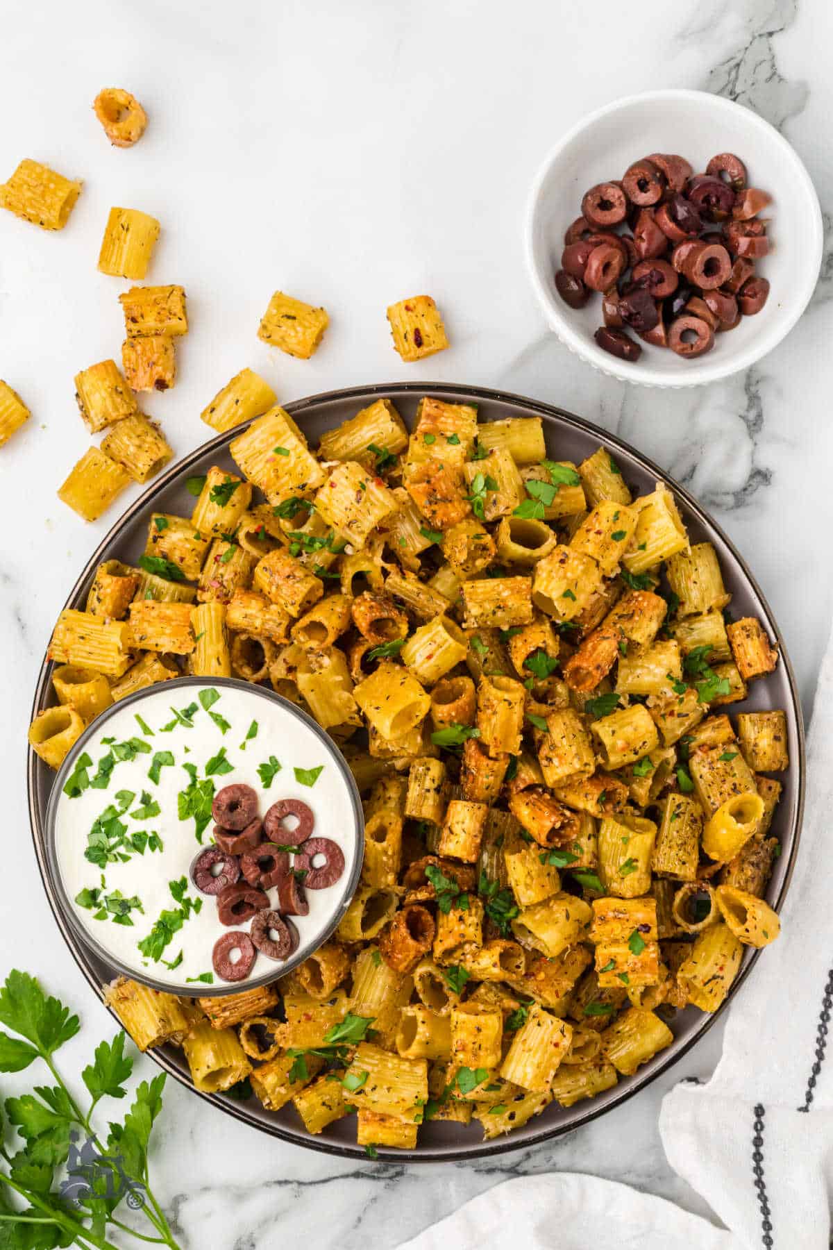 A large bowl filled with baked rigatoni pasta made into dippable chips and a creamy cheese dip with Kalamata olive on the side of the bowl. 