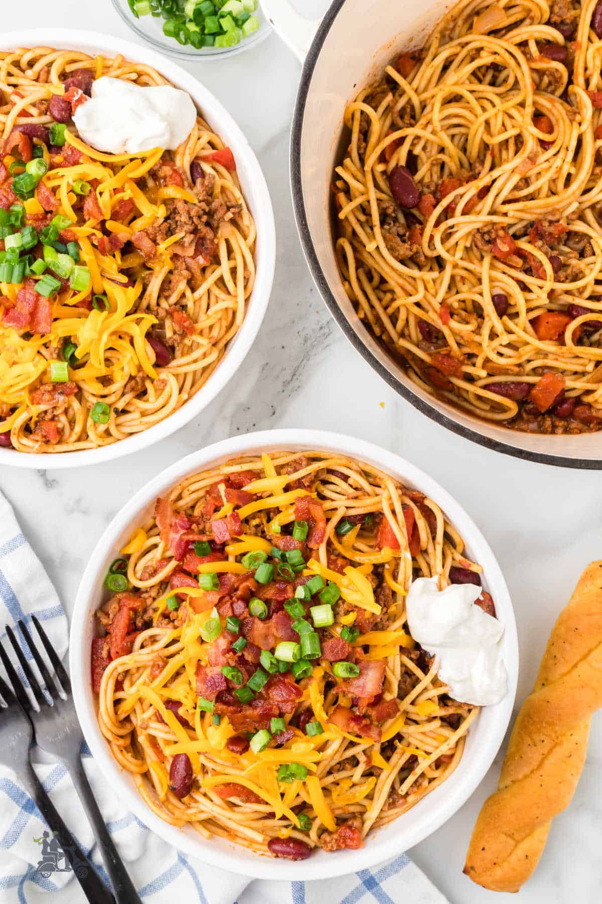 Cowboy Spaghetti topped with sour cream and chopped scallions in a white bowl with the cooking pot in the background.