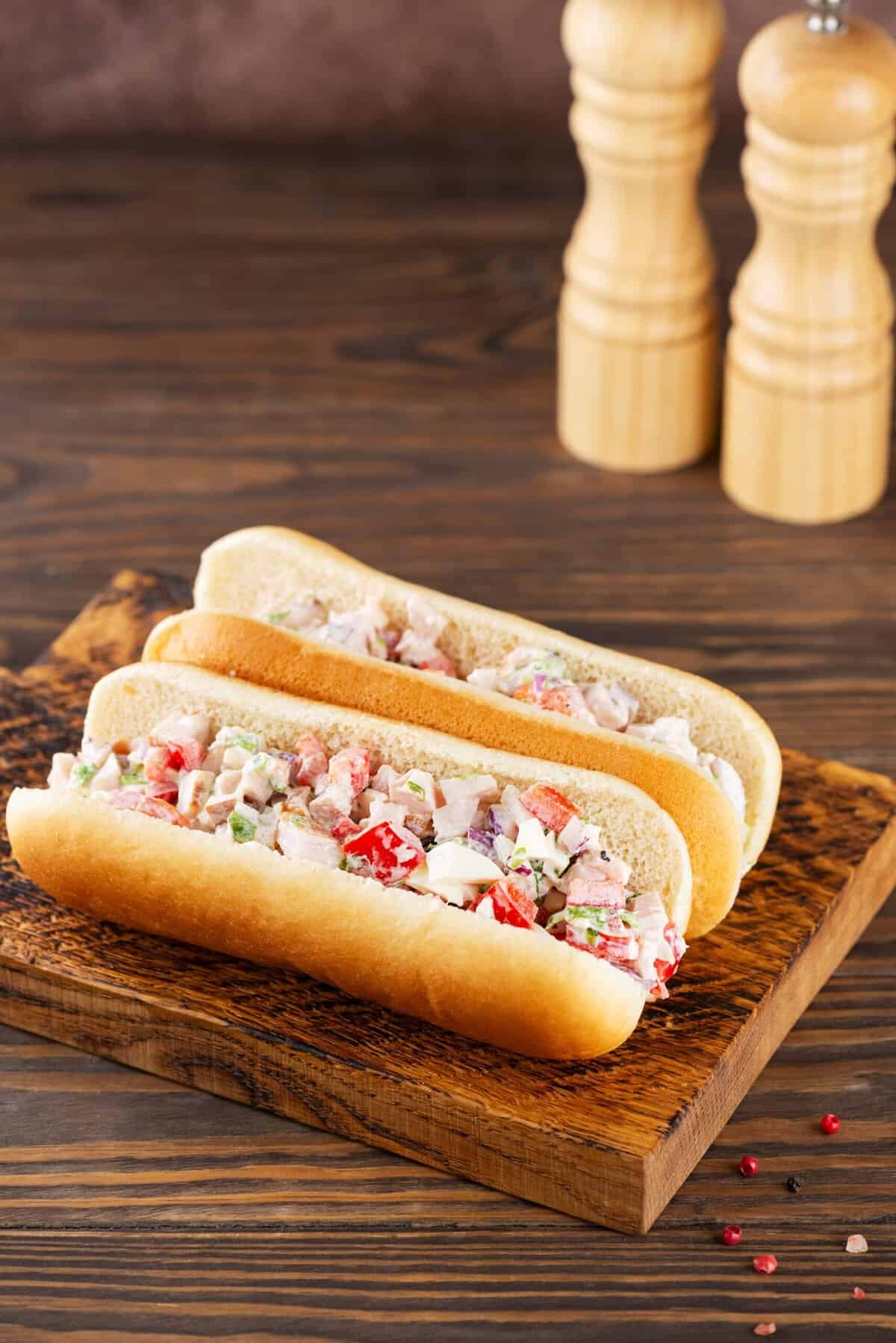 Image of an Italian chopped salad sandwich on a wooden board, showing layers of lettuce, meats, and cheese inside a hot dog bun. 