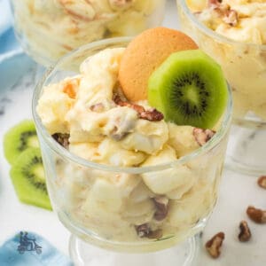 Banana pudding with marshmallows in a goblet, accompanied by vanilla wafers and kiwi in a glass.