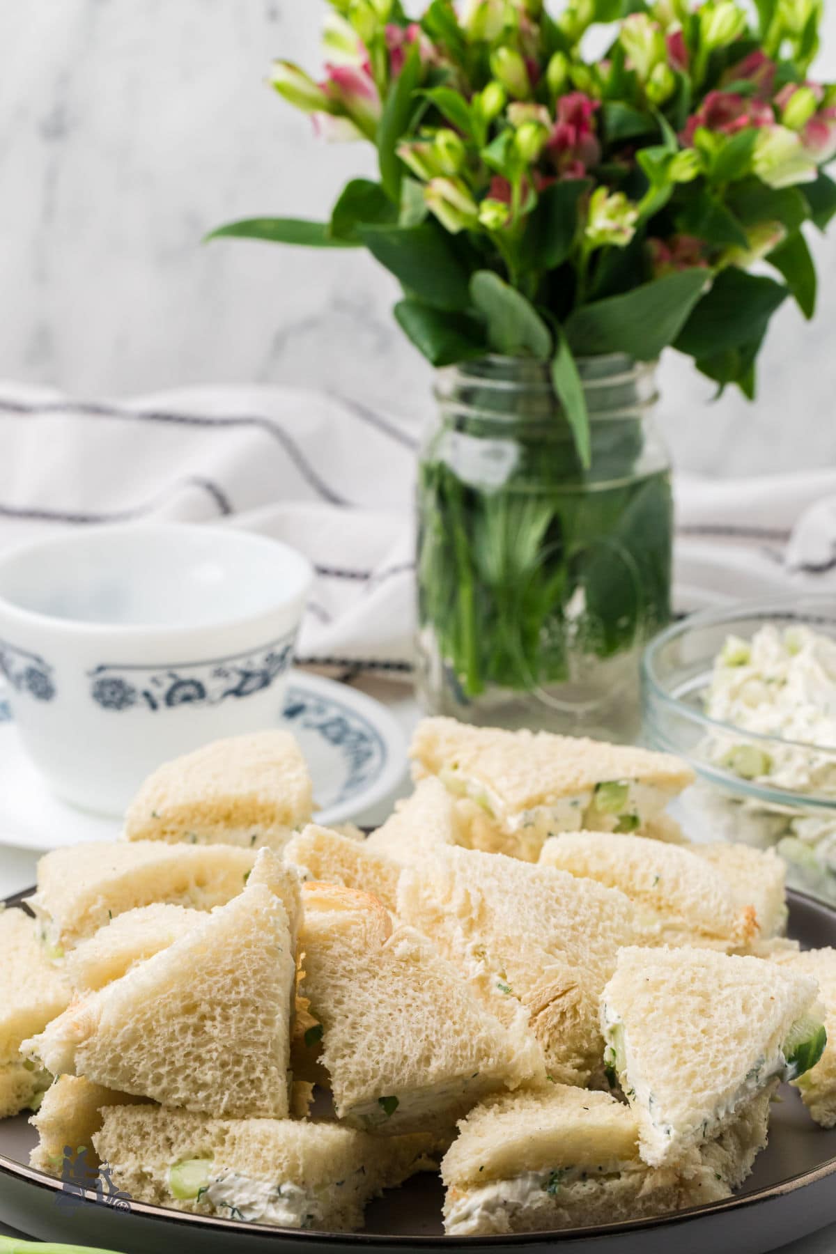 Cucumber tea sandwiches piled on a brown serving plate with a bouquet in the background. 