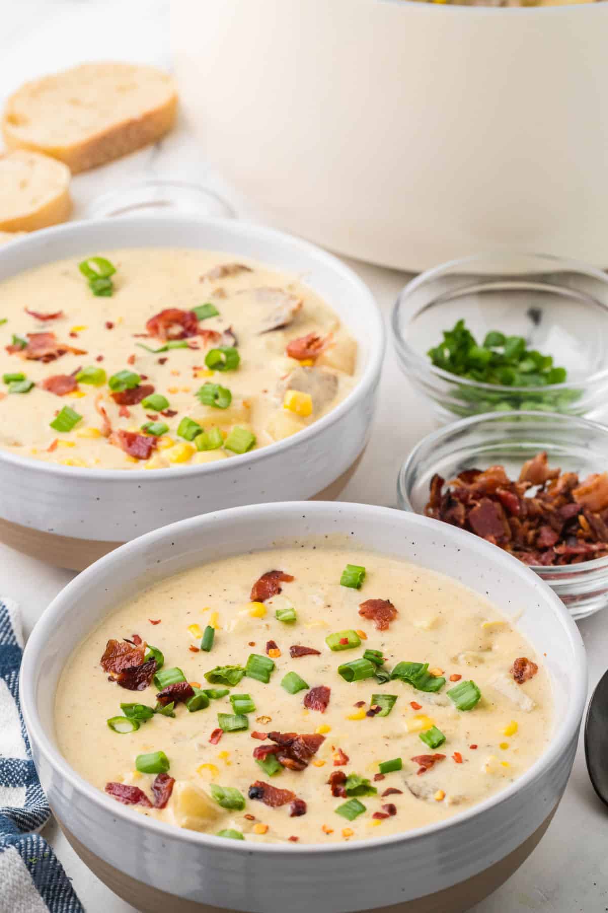 Creamy corn chowder with potatoes and spicy sausage in two bowls garnished with bacon, green onions, and red pepper flakes. 