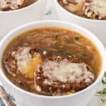 A white soup bowl filled with French Onion Soup recipe topped with a baguette slice sprinkled with melted Gruyère cheese.