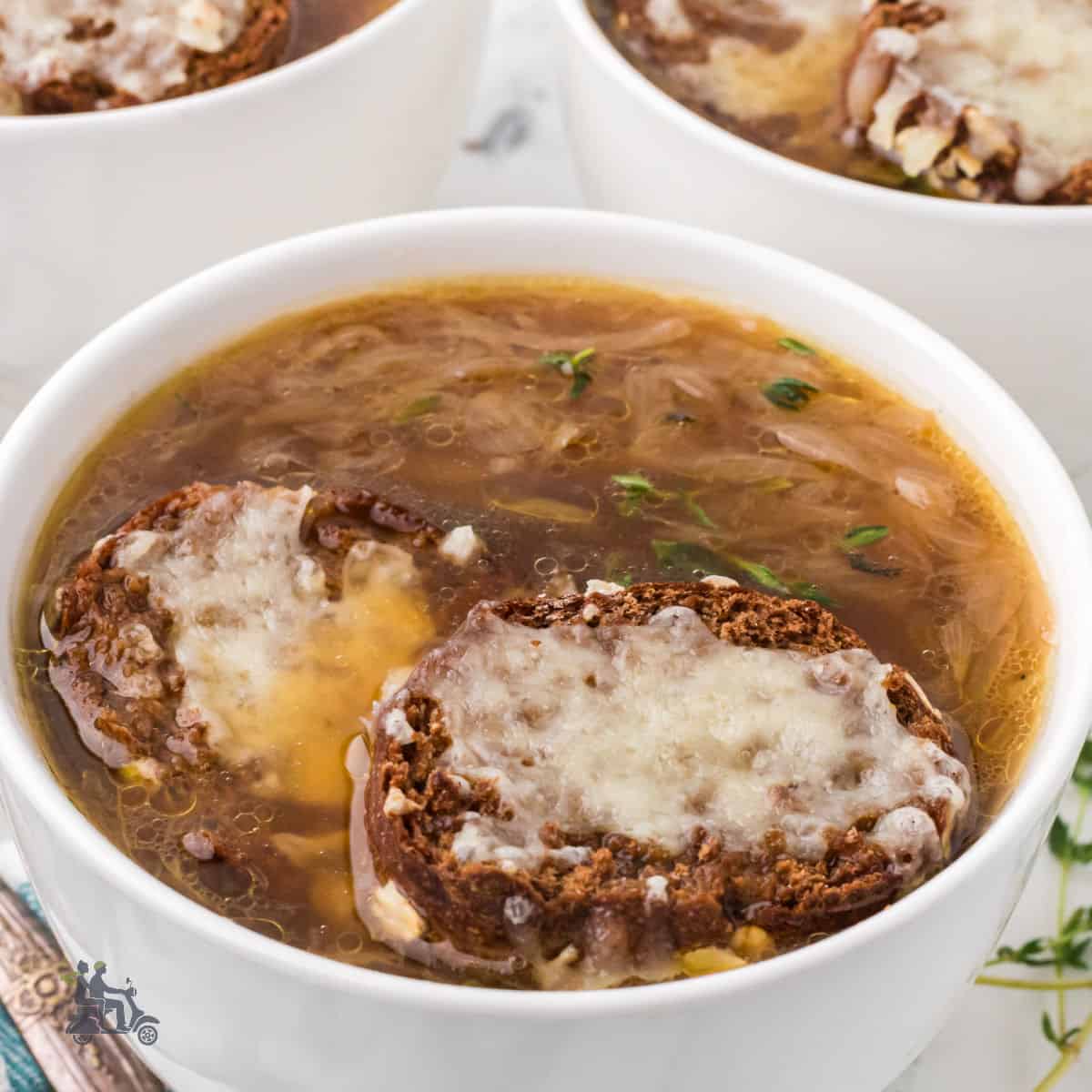A white soup bowl filled with French Onion Soup recipe topped with a baguette slice sprinkled with melted Gruyère cheese.