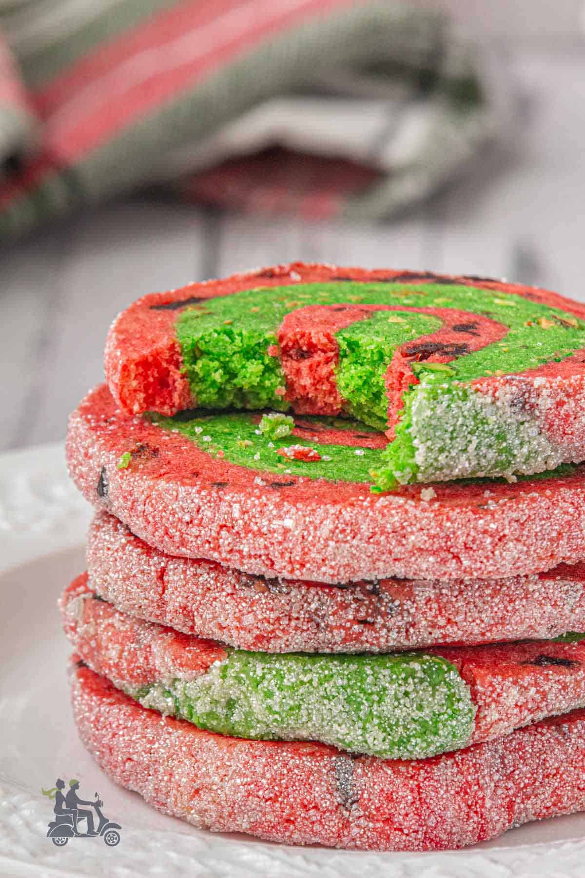 A stack of green and red cherry pistachio pinwheel cookies on a white plate with a bite taken out of the top cookie.