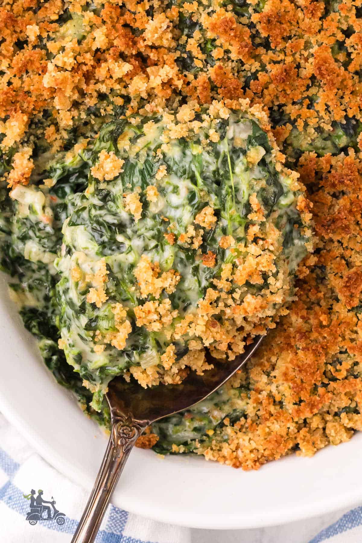 A spoon removes a serving of creamed spinach covered with breadcrumbs from the serving dish.