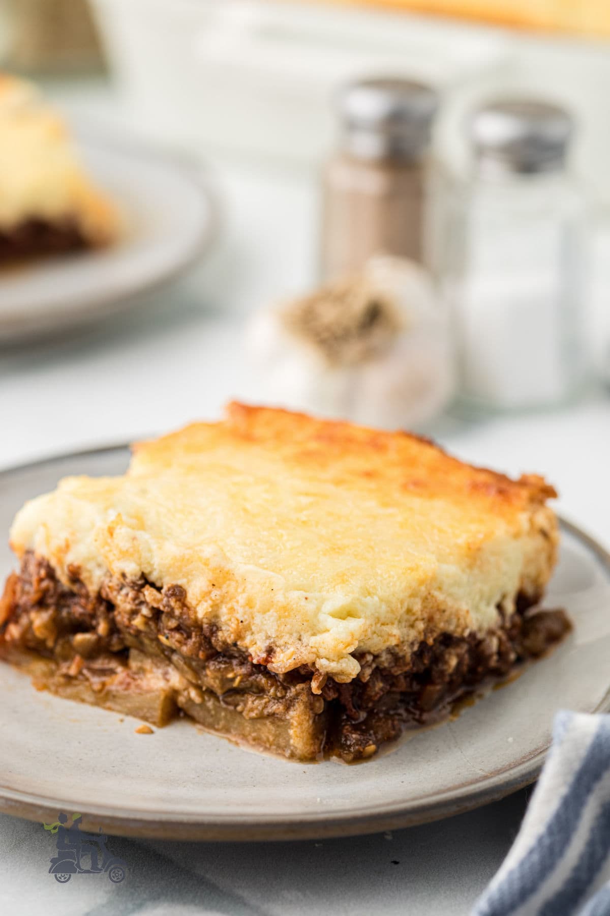 A homemade moussaka serving on a plate with a salt and pepper shaker in the background. 