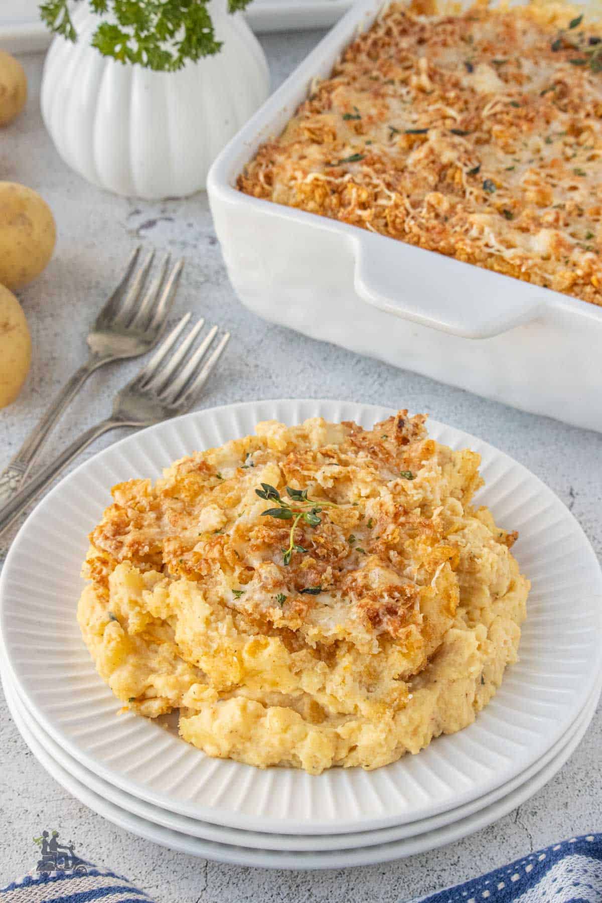 Make-ahead cheesy mashed potatoes baked in a casserole dish with a helping on a white saucer.